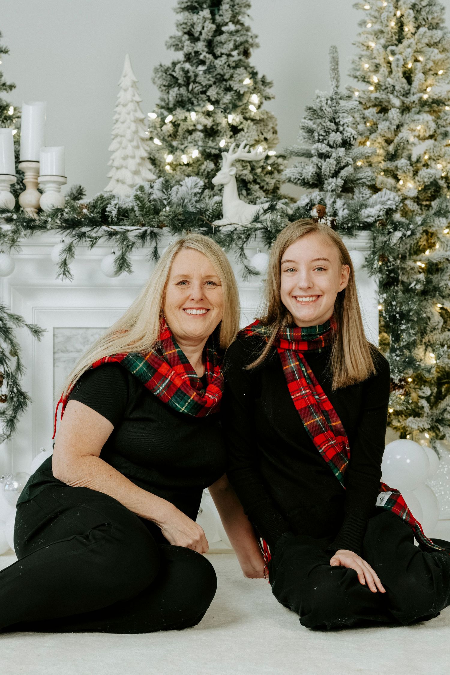 Two people in black outfits and plaid scarves pose on floor in front of decorated Christmas trees.