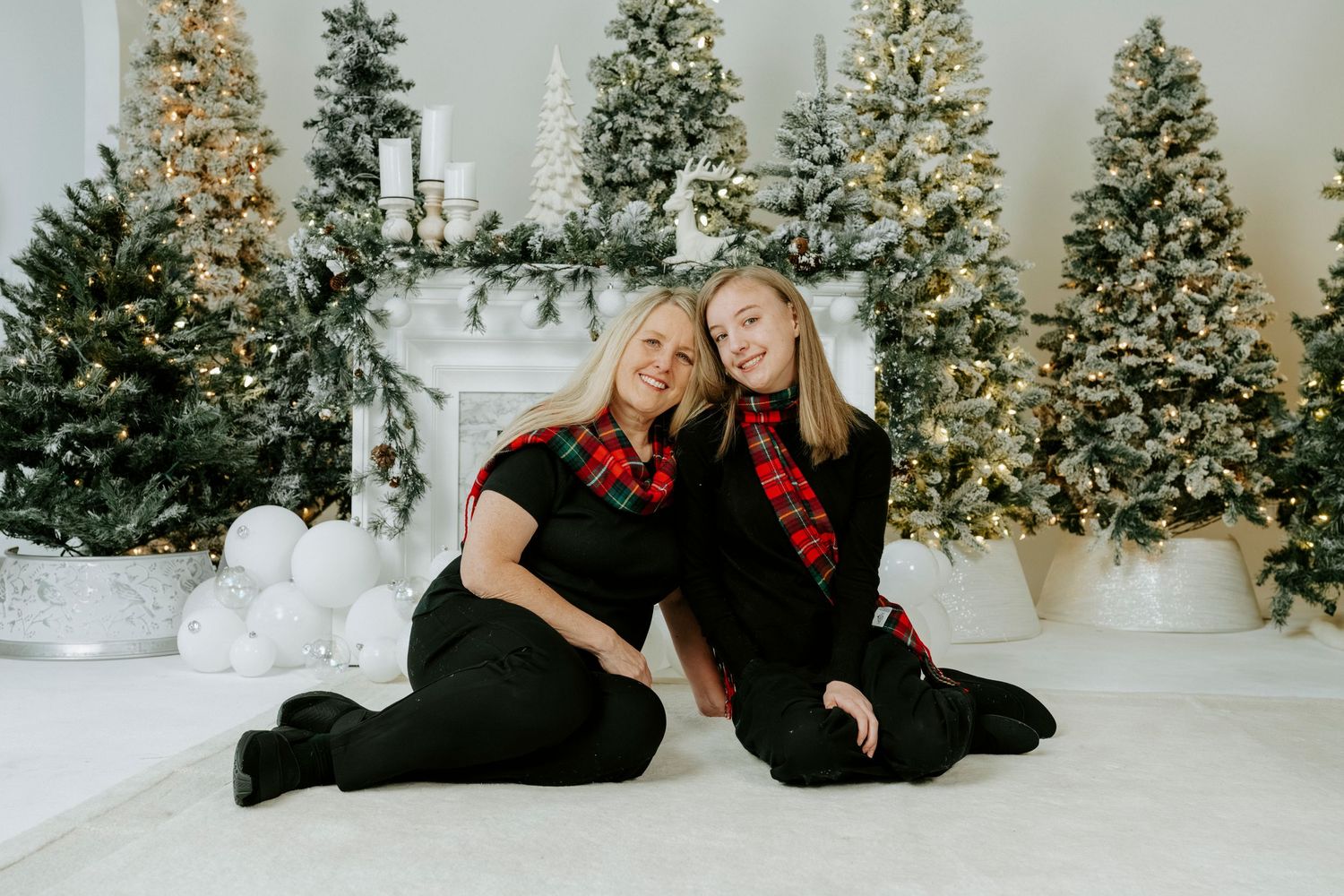 Two people in coordinating black and red outfits sit on white floor with illuminated Christmas trees behind them.