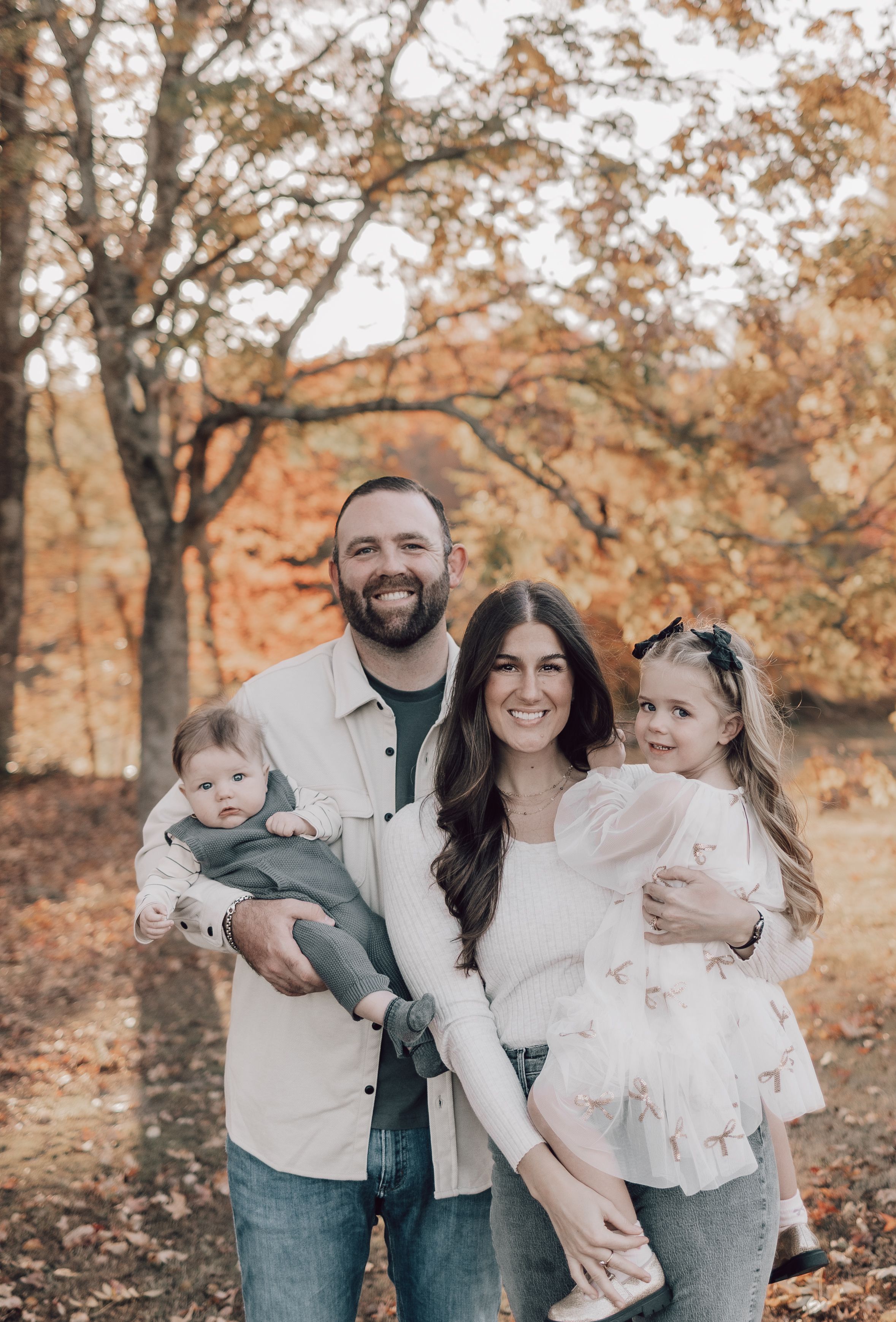 A family in coordinating white and denim outfits poses together during an autumn photoshoot with golden leaves in background.