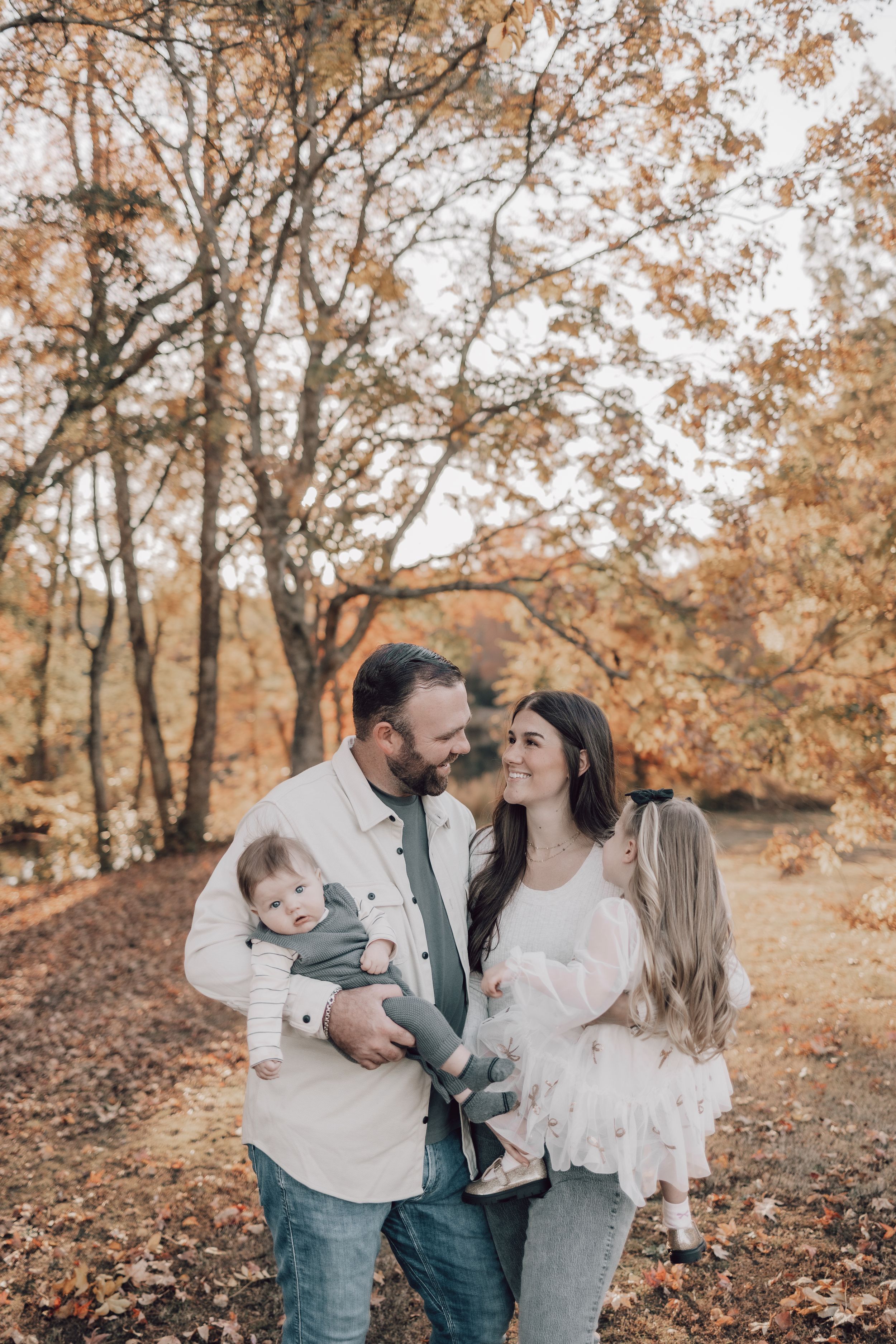 Family in coordinating white and sage outfits share loving moments among autumn trees with fallen leaves.