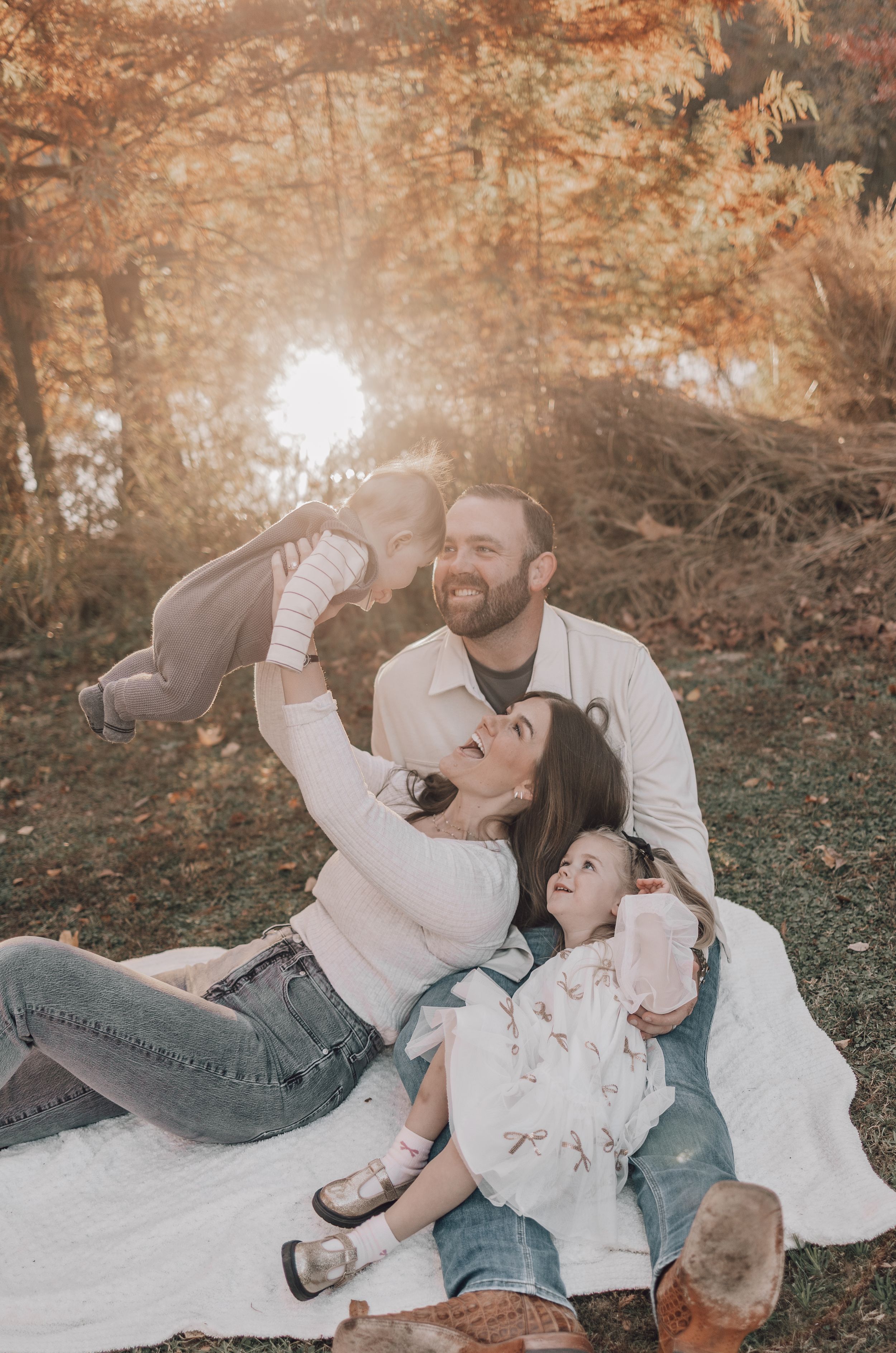 A family sits together on a blanket in an autumn forest setting with golden sunlight filtering through the trees.