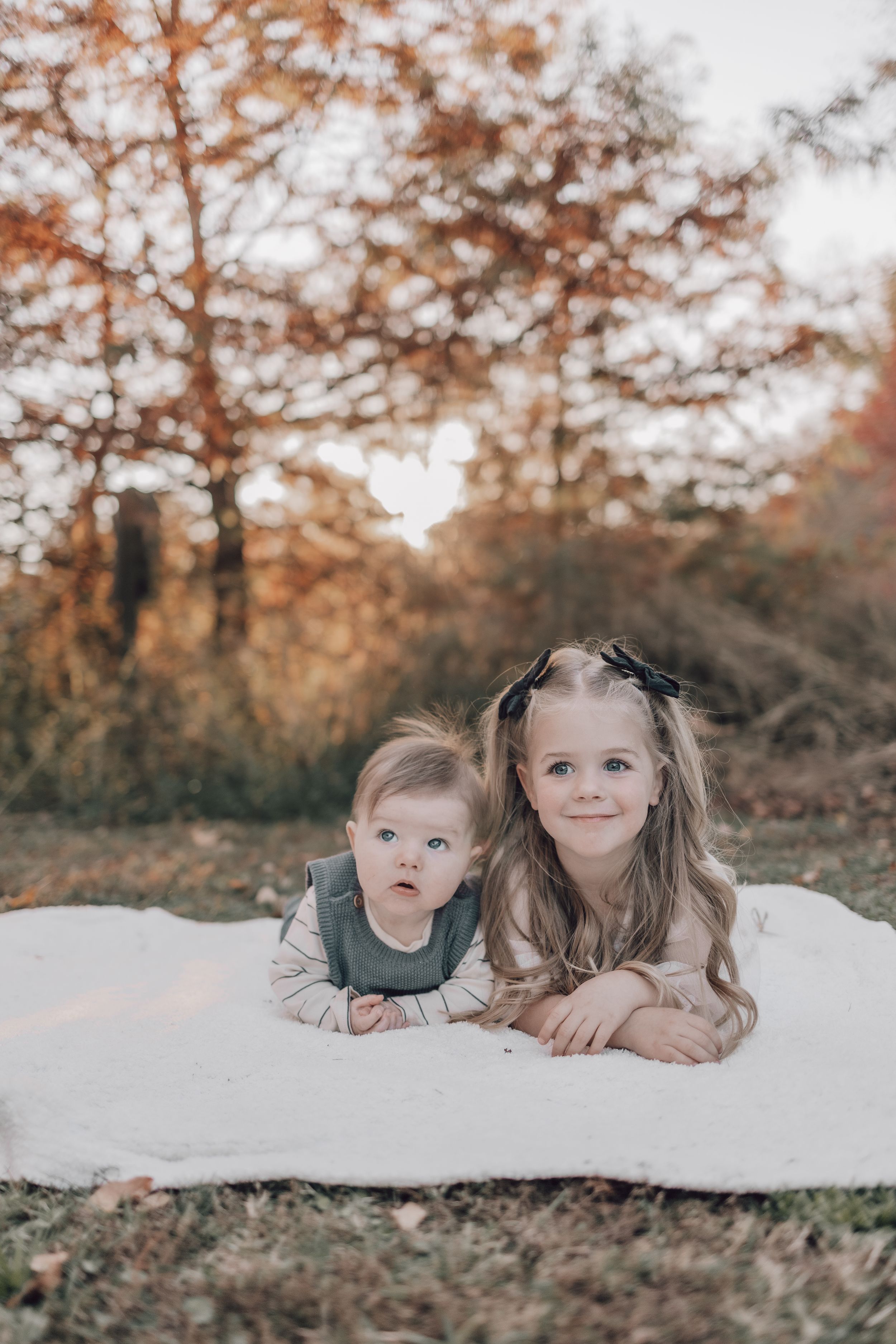 Two children lying on a white blanket outdoors during fall with autumn trees in the background.