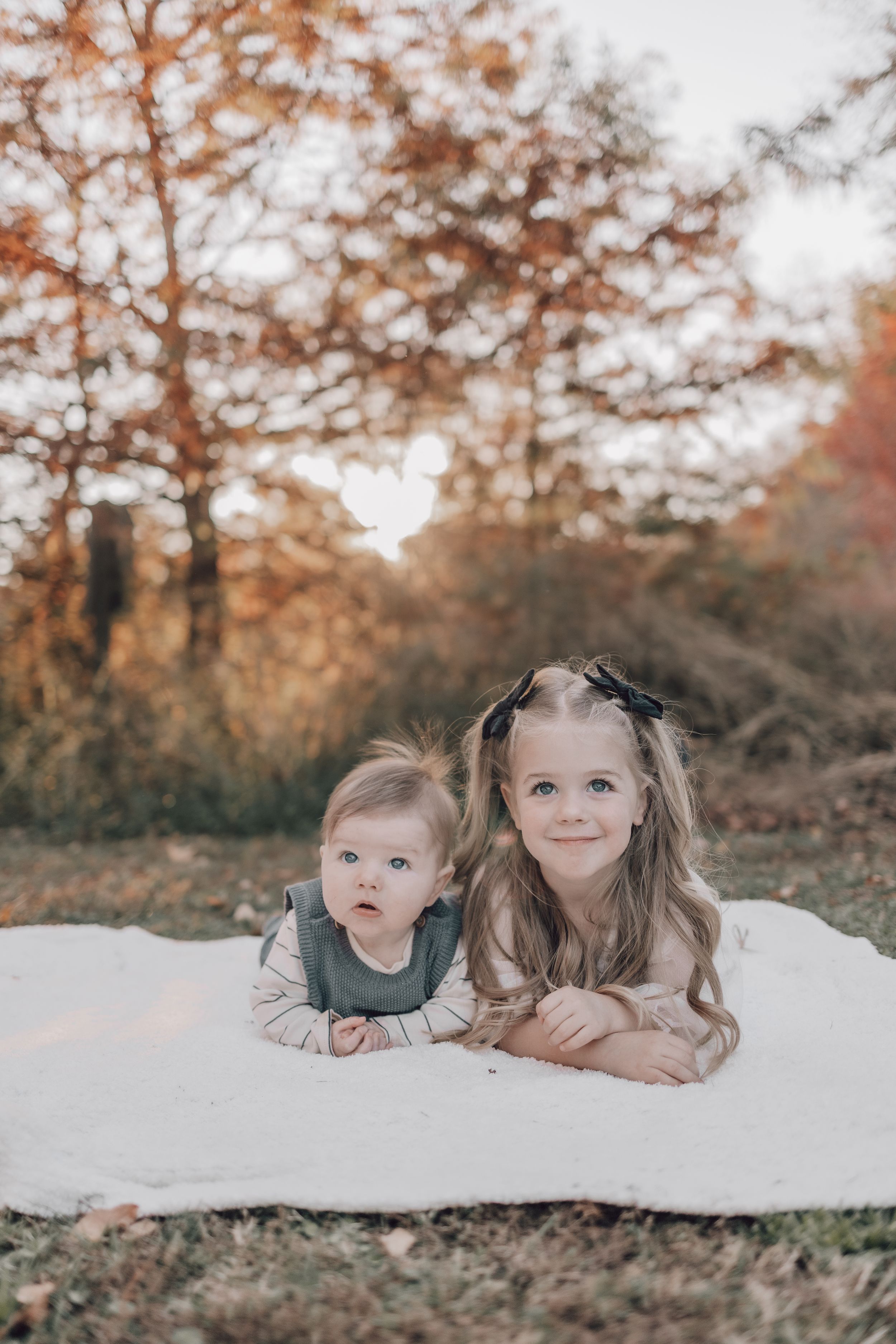 Two siblings posing on white blanket during autumn photoshoot with fall foliage in background.