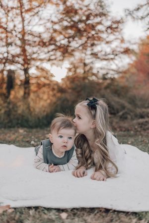 Sweet autumn portrait of siblings lying on white blanket with fall trees in background.