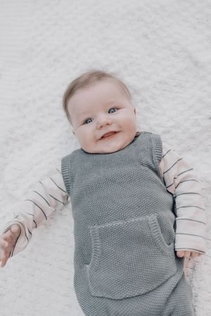 Baby wearing grey knit vest and striped sleeves lying on white background looking up with a smile.