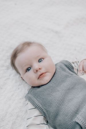 Baby wearing a gray knit sweater lies on white fabric while looking upward with bright blue eyes.