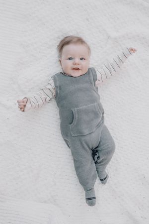 Baby in grey knit romper and striped sleeves lying on white background with arms spread out.