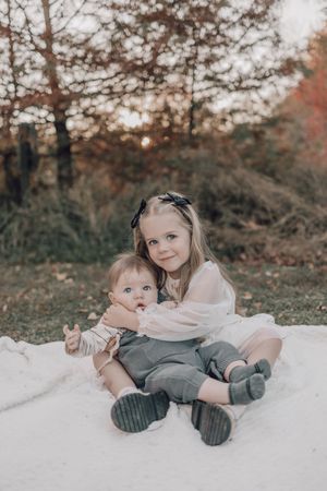 Two children sitting together on a white blanket during an autumn photoshoot outdoors.