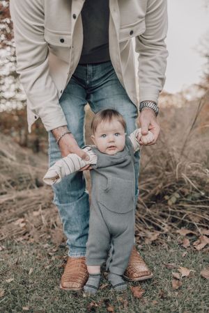 Close-up of small child learning to walk while holding parent's hands on grass.