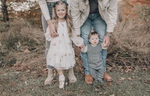 A family in casual attire stands together holding hands outdoors during autumn with a rustic backdrop.