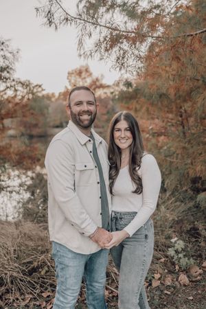 A couple poses together in casual attire against a backdrop of fall foliage near a waterfront.