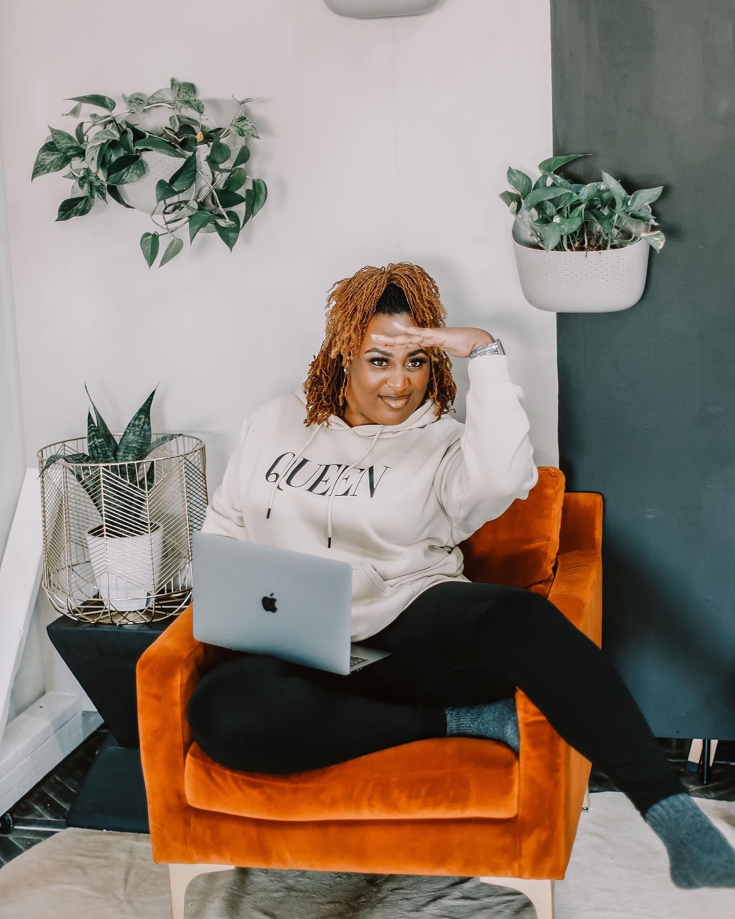 Someone working on a laptop while relaxing on an orange velvet couch with indoor plants in the background.