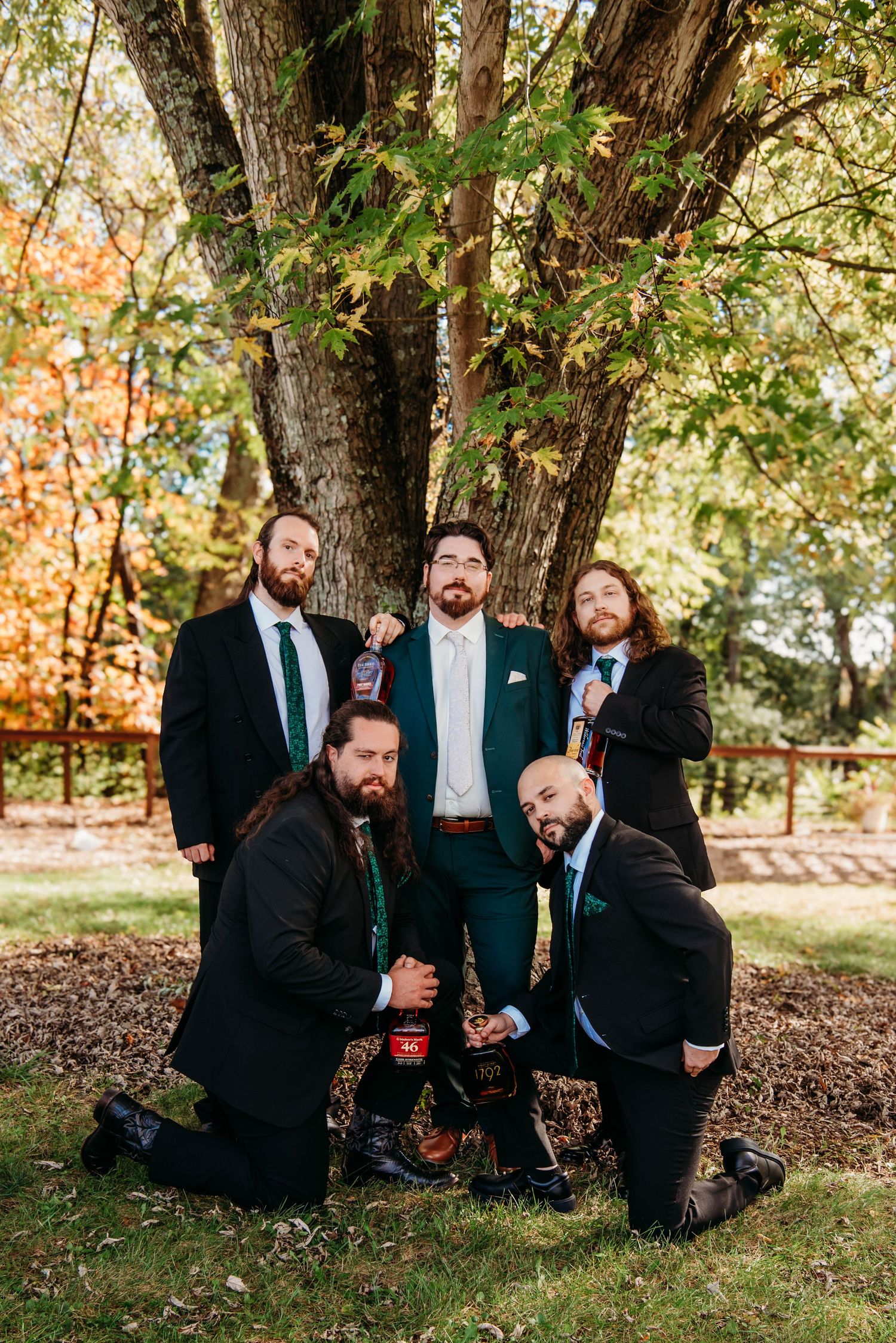 Groomsmen in dark suits share laughs while sitting together under autumn trees at an outdoor wedding.