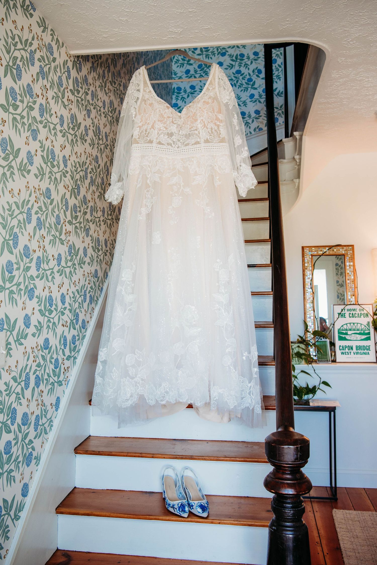 A delicate white lace wedding dress hangs on display with blue shoes on wooden stairs.
