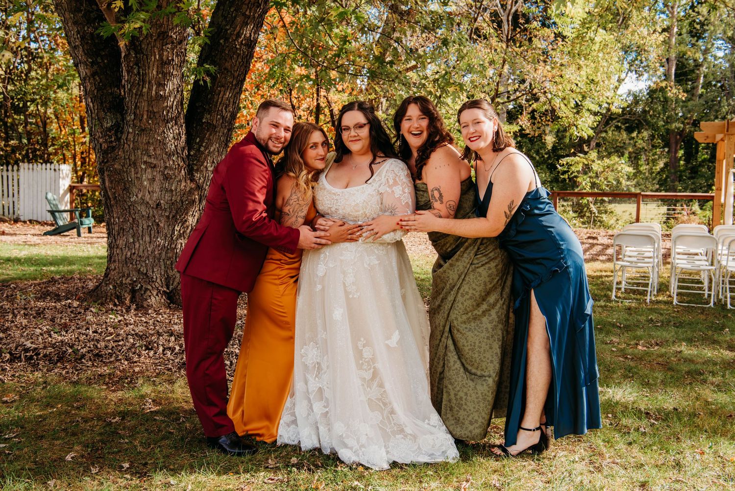 Wedding group poses together outdoors under large oak trees on a sunny autumn day.