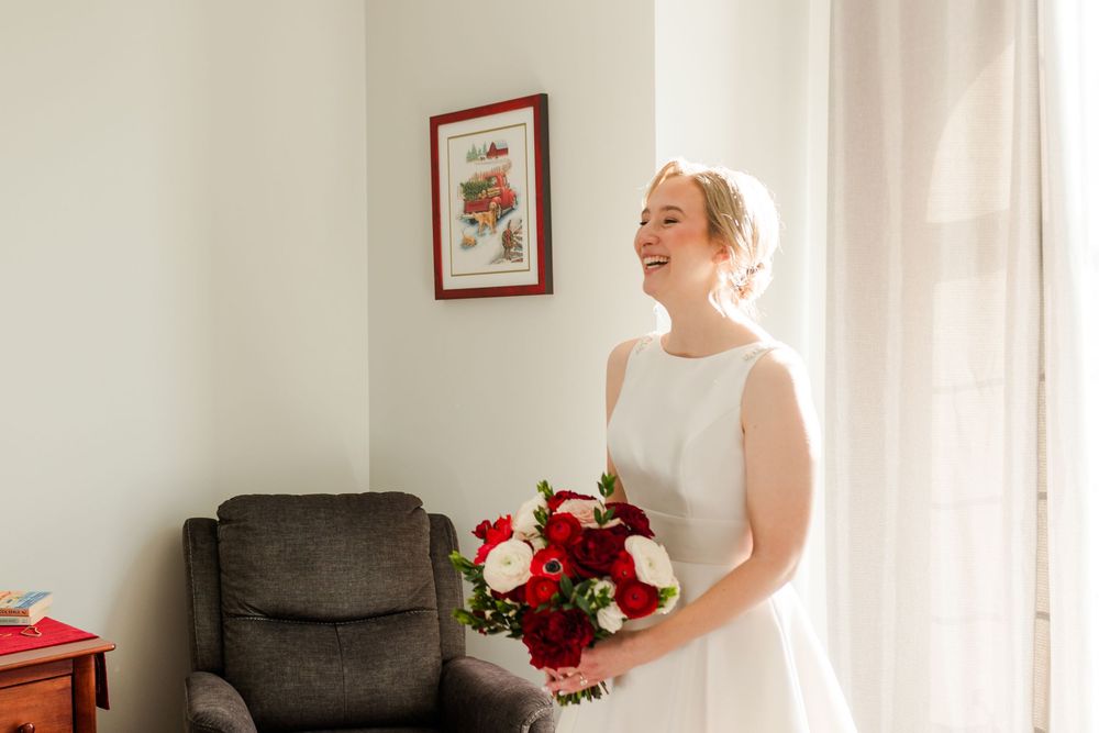 A wedding party poses together in a bright room with white curtains and red floral bouquets.