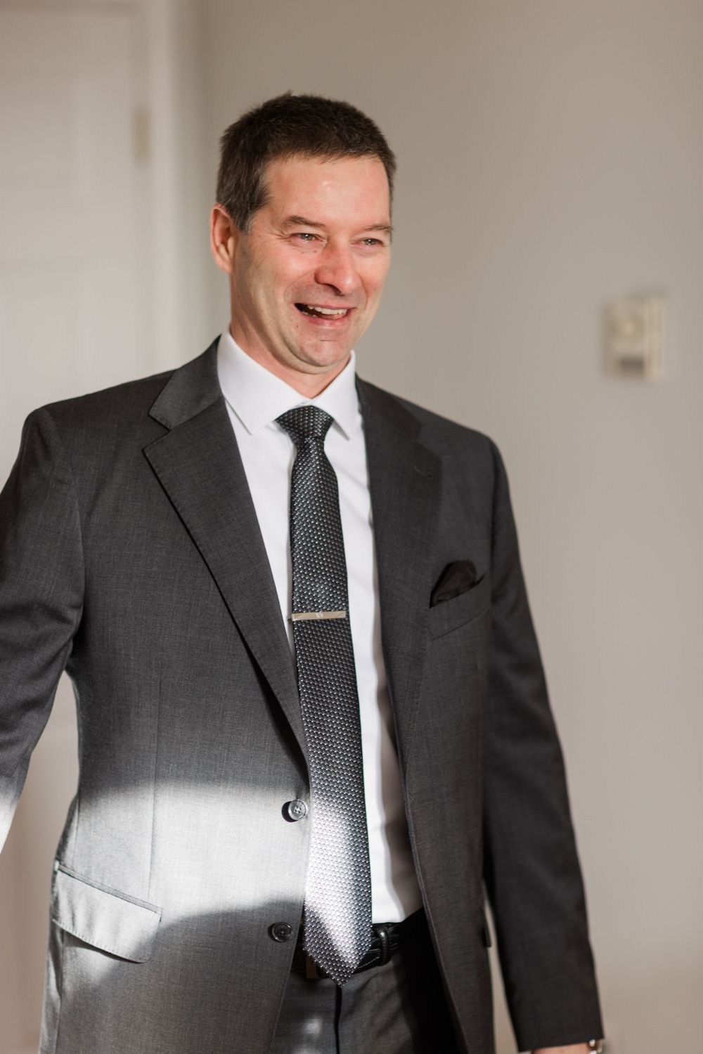 A person wearing a dark grey suit and tie laughs joyfully while standing in a bright indoor setting.
