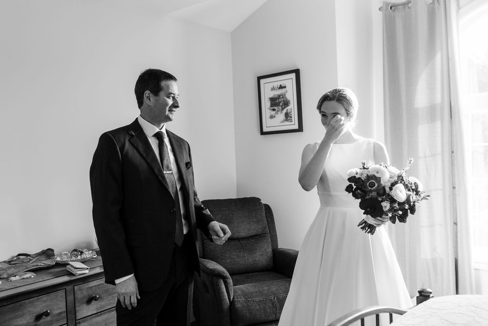 A black and white wedding photo showing a couple in a bright interior room with a leather sofa and window.