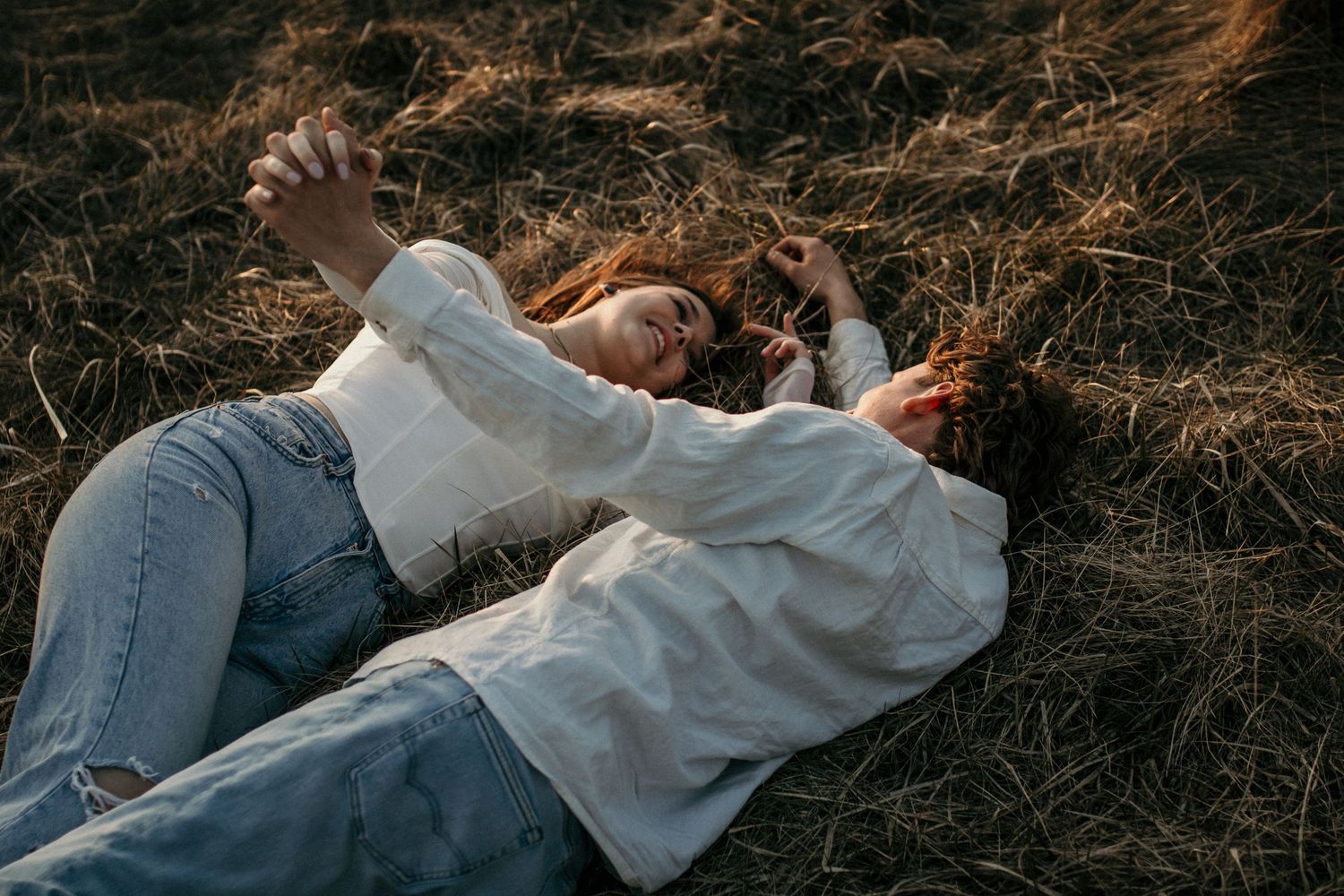 A person lying in dry grass wearing casual denim and white clothing during an outdoor photo session.