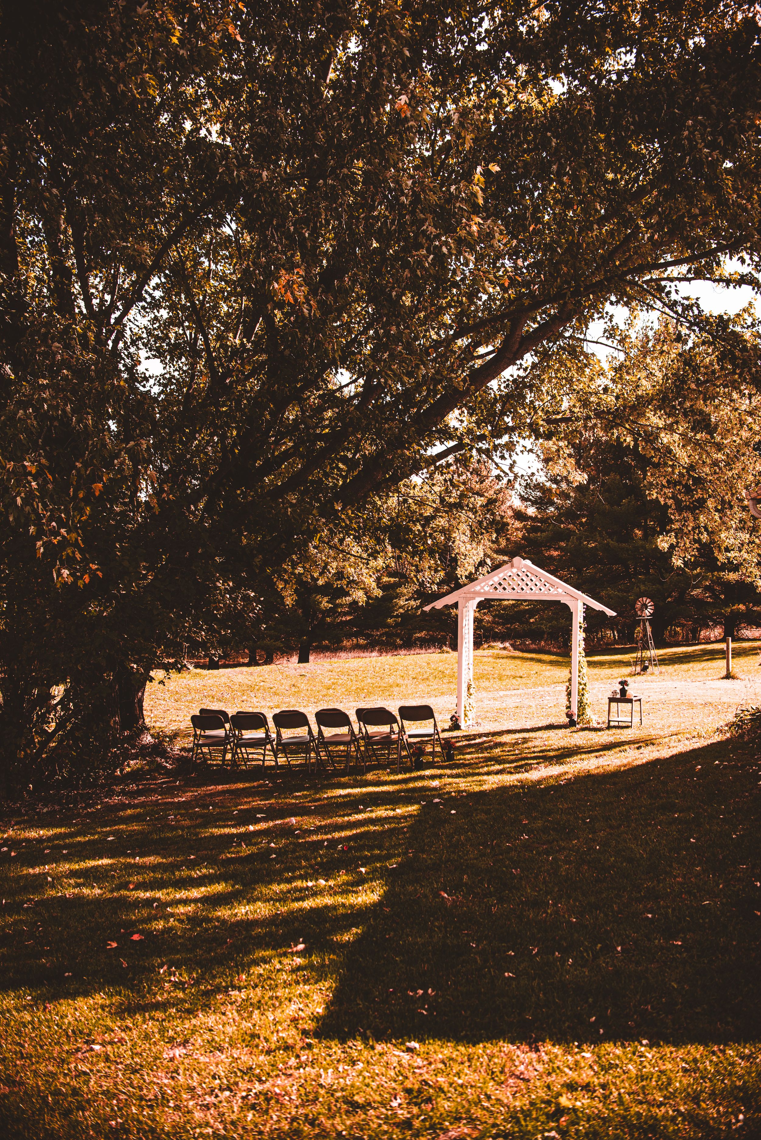 Rustic outdoor wedding venue with wooden gazebo and chairs under autumn trees.
