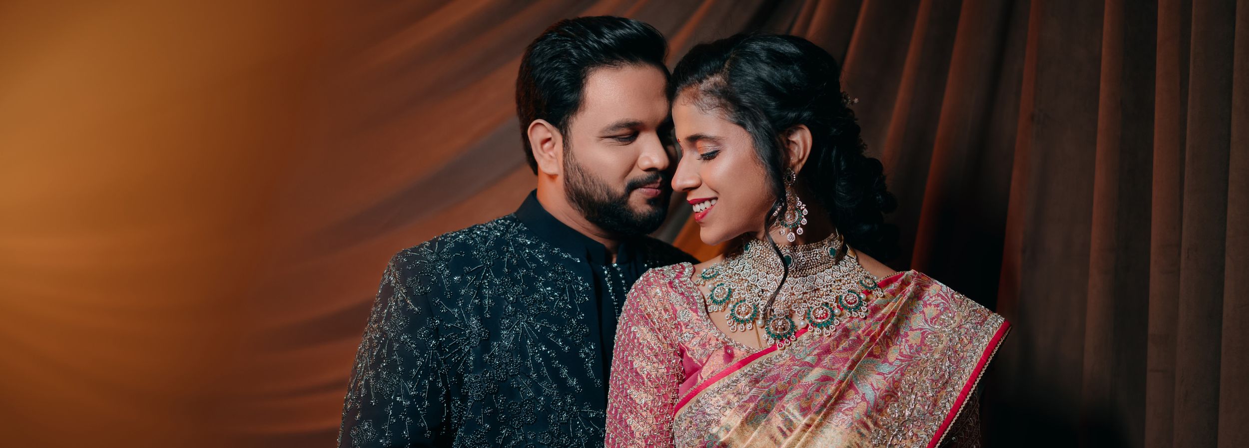 Intimate portrait of couple in ornate traditional Indian wedding attire against warm-toned backdrop.