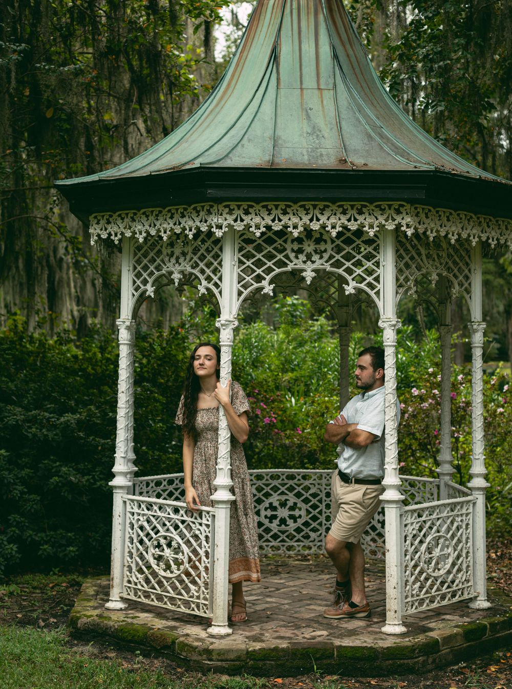 A couple poses in a vintage white gazebo surrounded by Spanish moss and trees.
