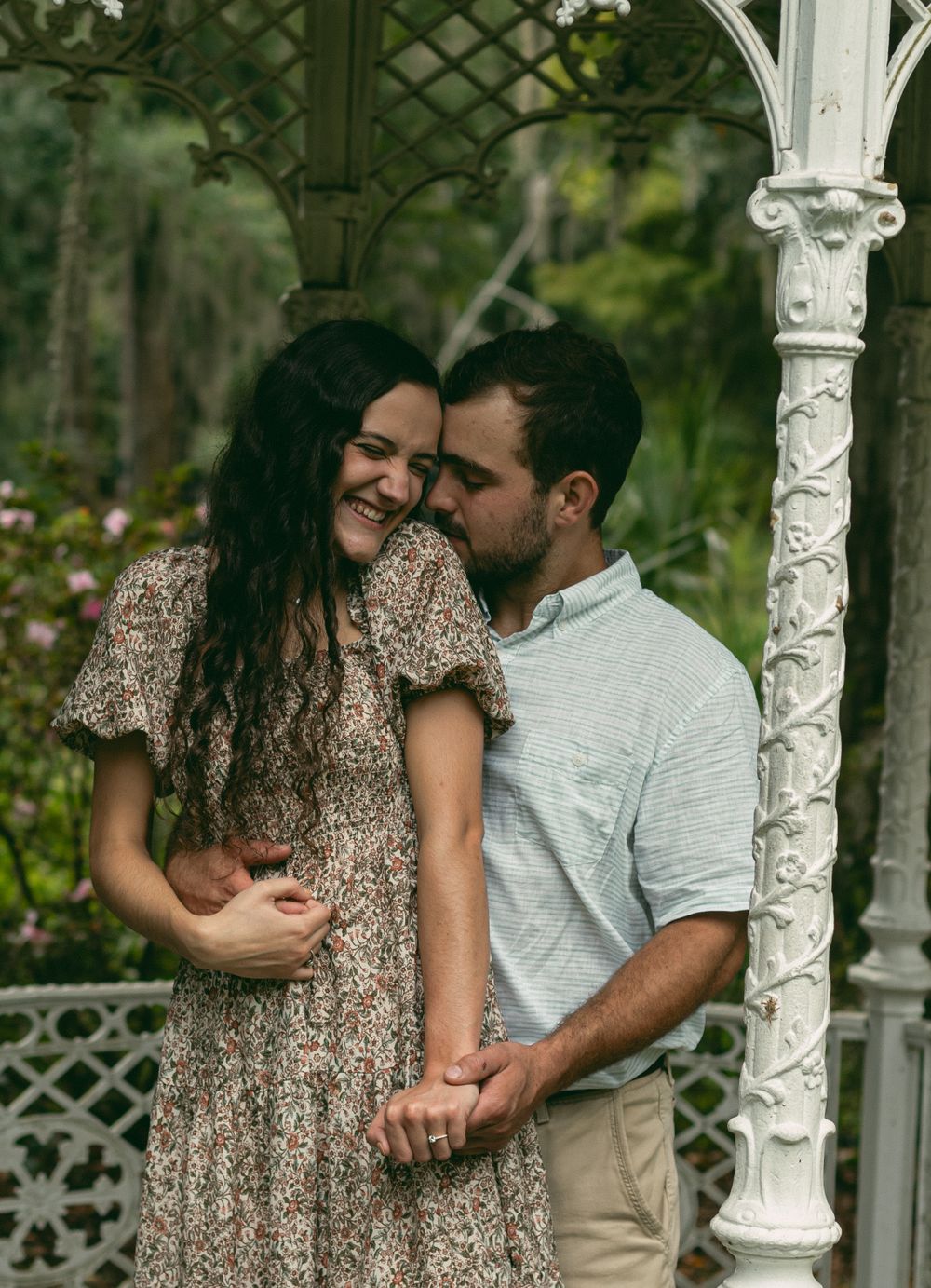 A couple shares a tender embrace in an ornate white gazebo surrounded by greenery.