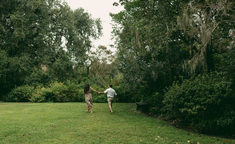 Two people run playfully across a lush green lawn towards a wooded area.