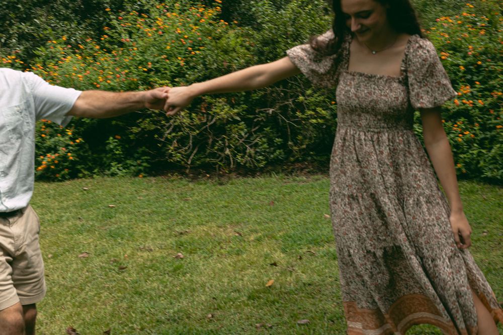 Two people hold hands and dance in a grassy outdoor area surrounded by greenery.