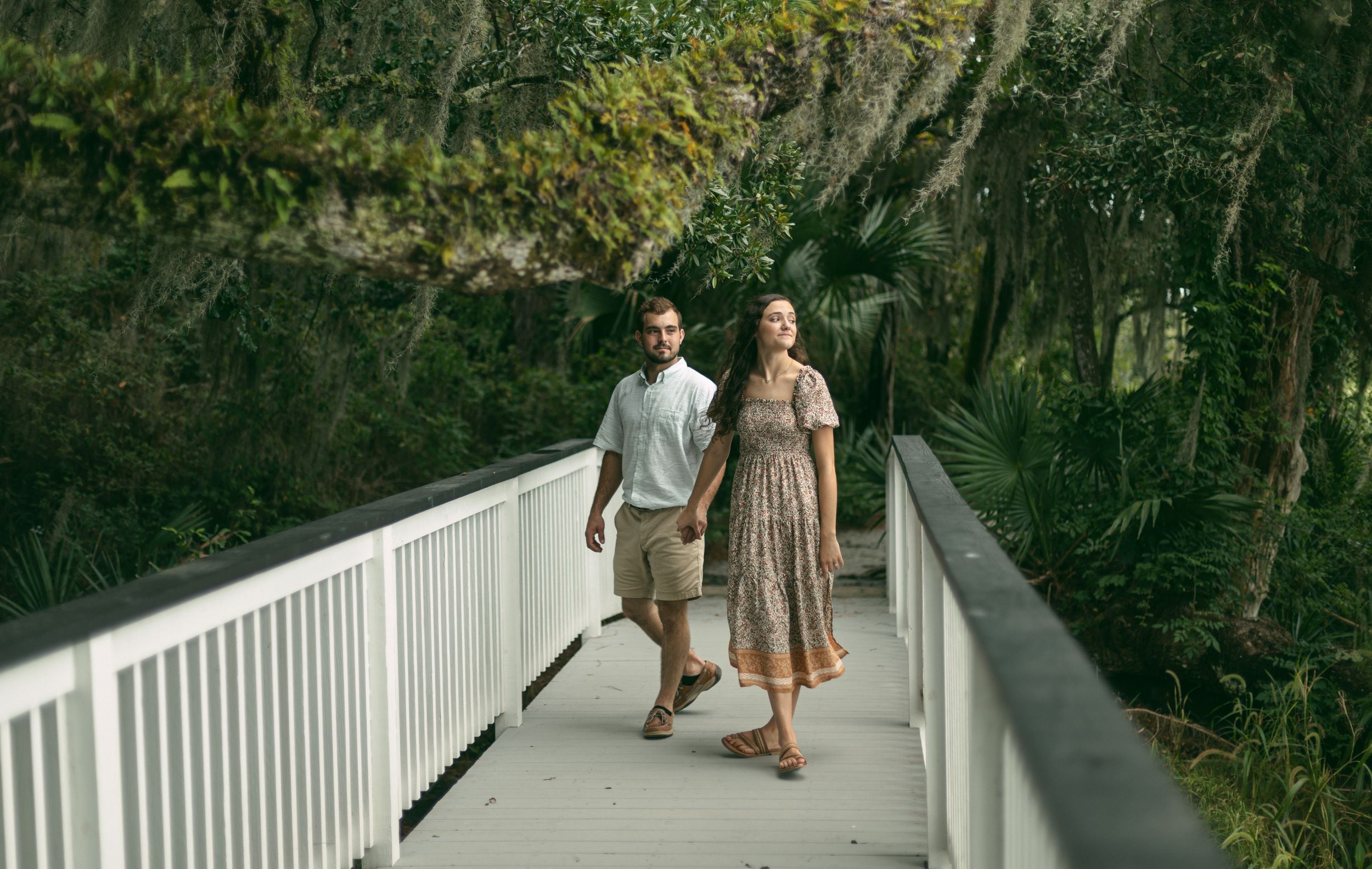 A romantic couple strolls together on a white boardwalk surrounded by lush greenery and trees.