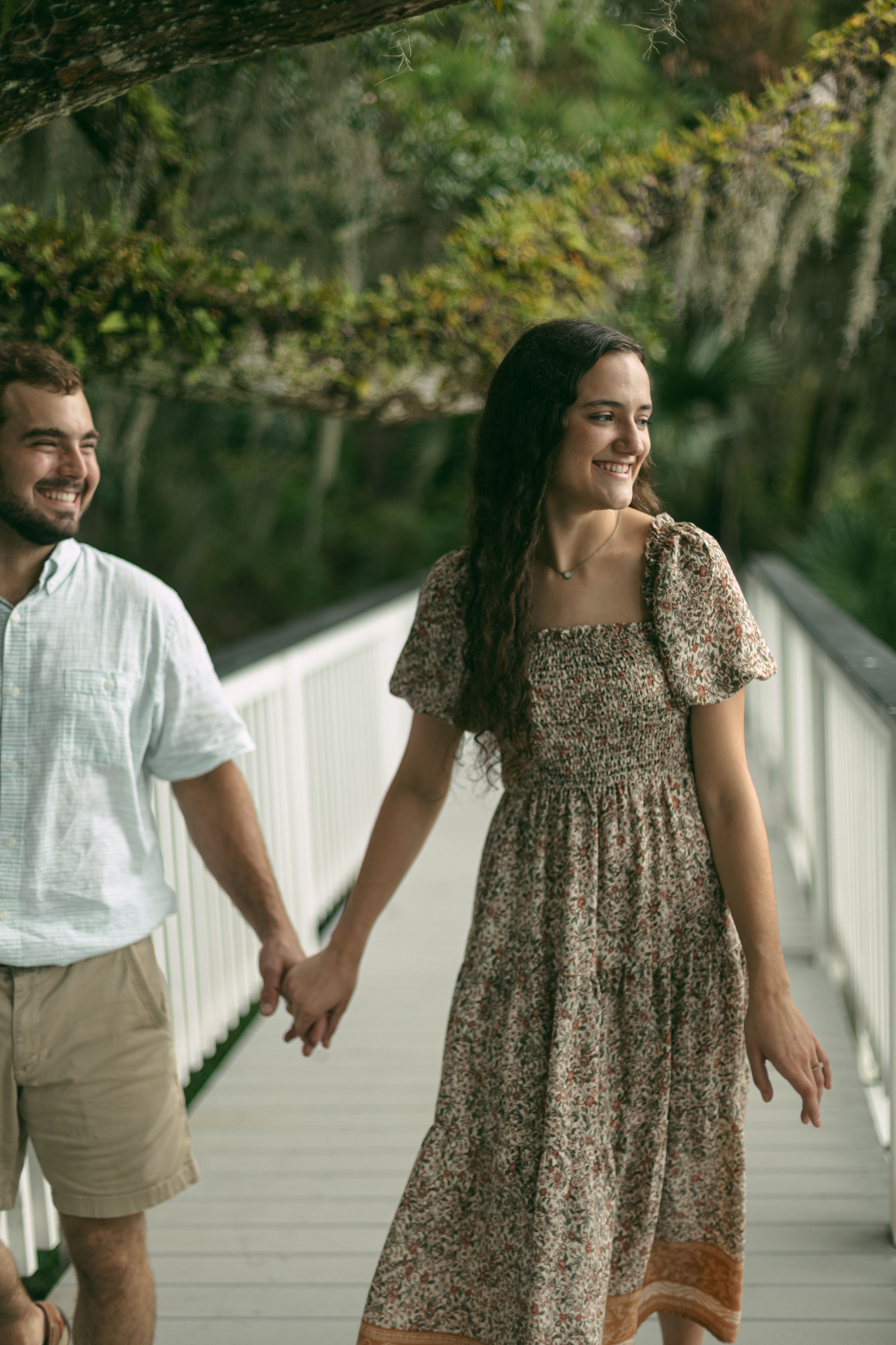 A joyful couple holds hands while walking on a raised white walkway surrounded by trees.