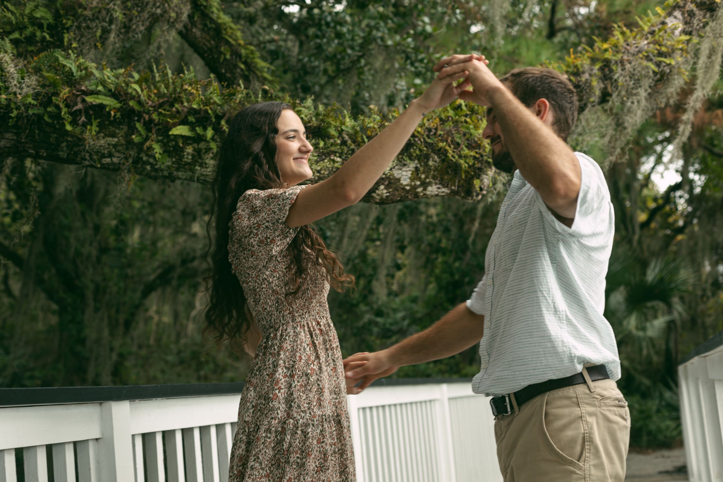 Two people dancing playfully on a white boardwalk surrounded by green foliage.