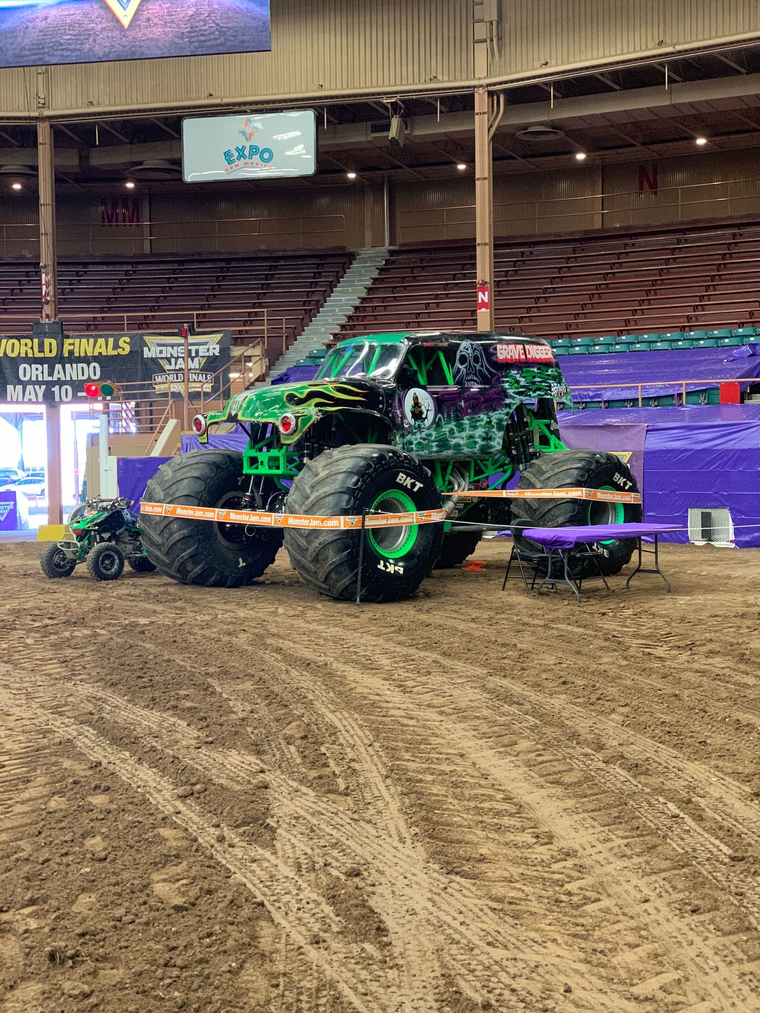 Large green monster truck displayed inside an indoor arena with dirt floor.