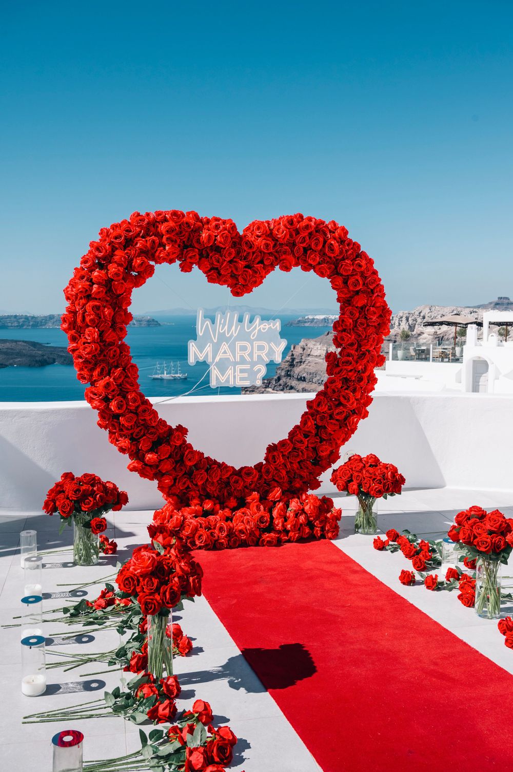 Red rose heart-shaped arch with 'Marry Me' sign and red carpet against Santorini's ocean view.