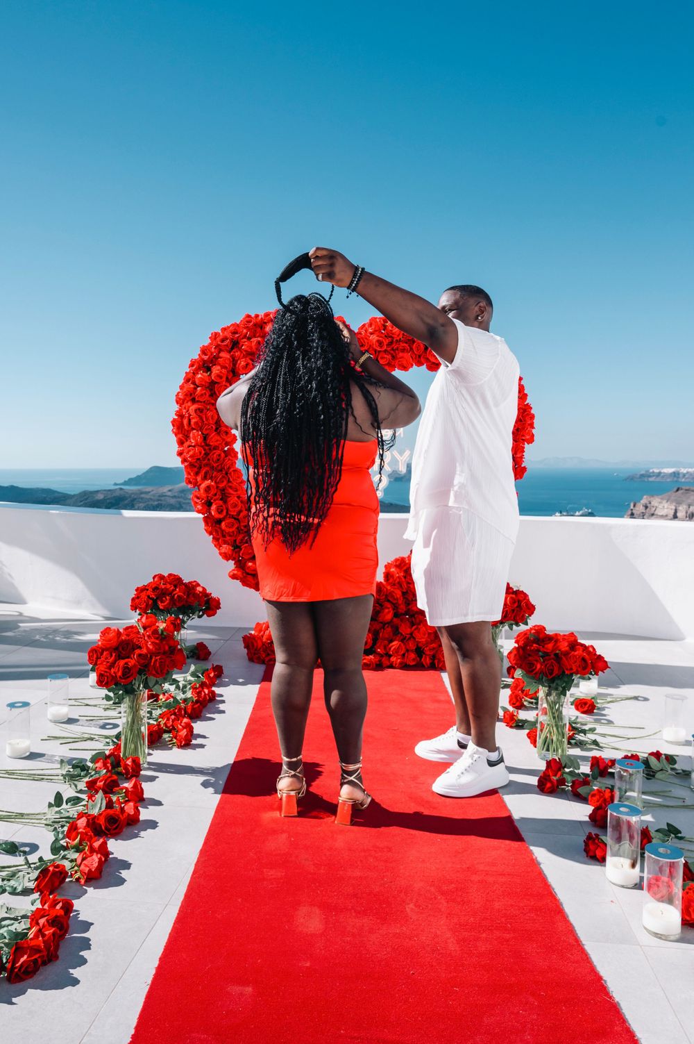 A couple stands on a red carpet under a floral heart arch during a proposal in Santorini.