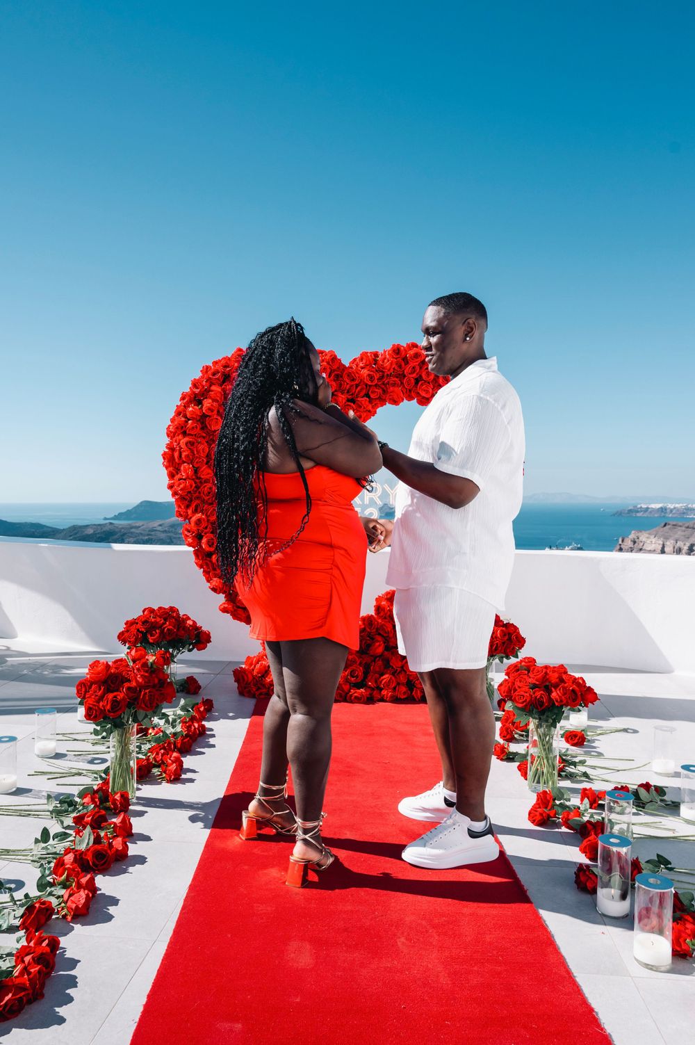 A romantic proposal moment under a red rose heart arch with ocean views in Santorini.