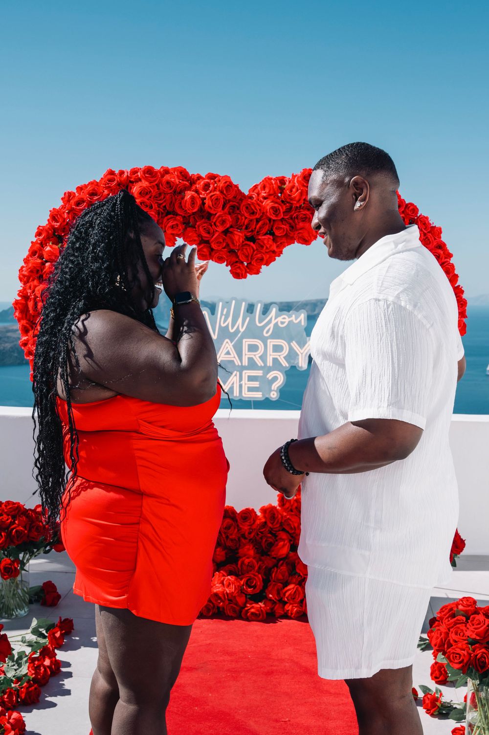 A couple shares an intimate moment during a marriage proposal under a red floral heart arch.