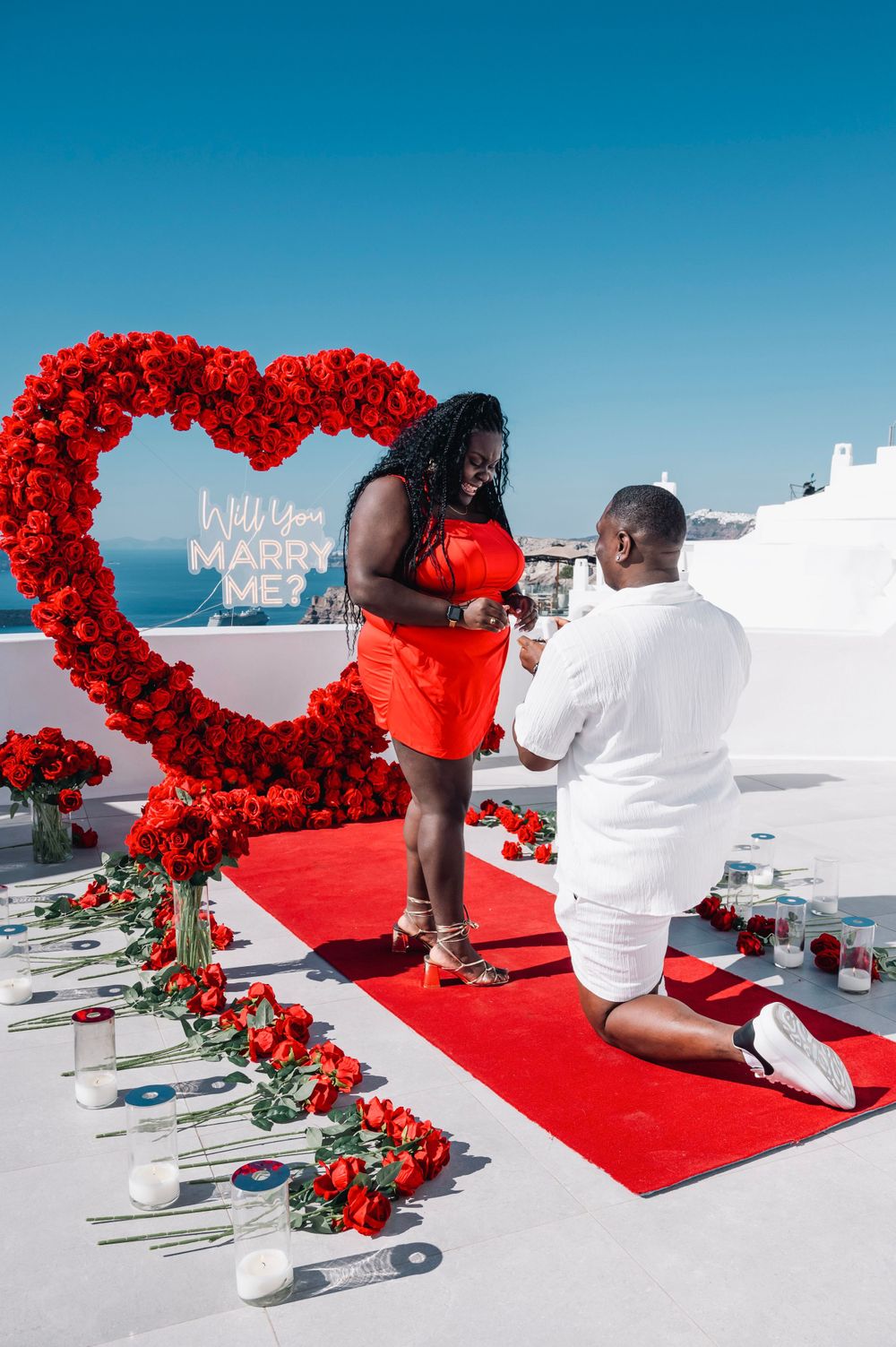A romantic proposal scene with someone kneeling under a red rose heart arch in Santorini.