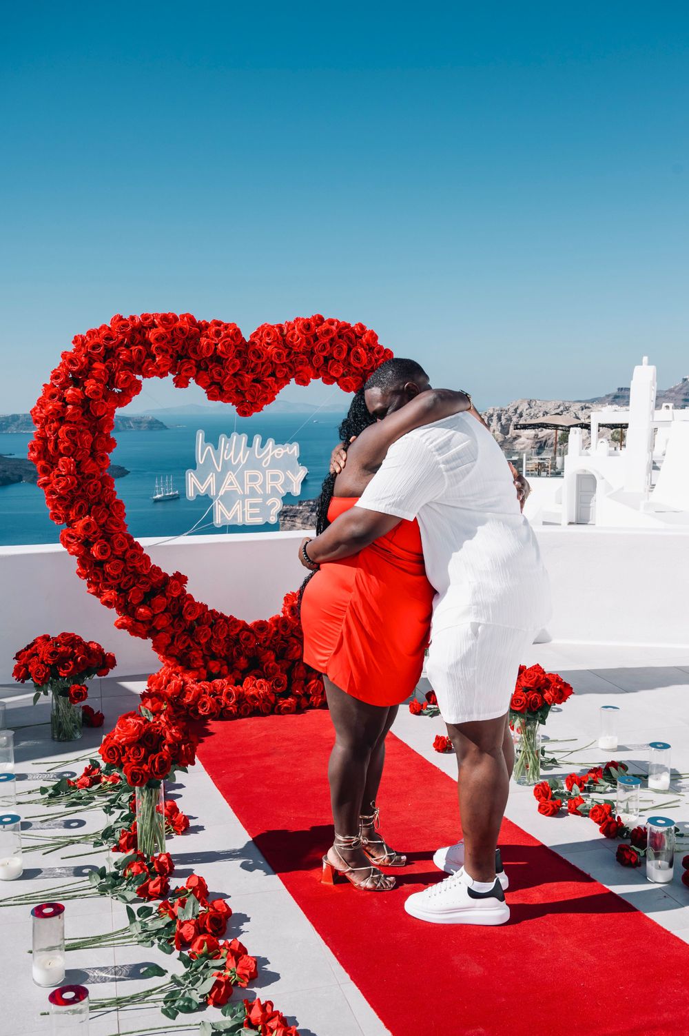A couple embraces after a proposal under a red heart-shaped flower arch in Santorini.