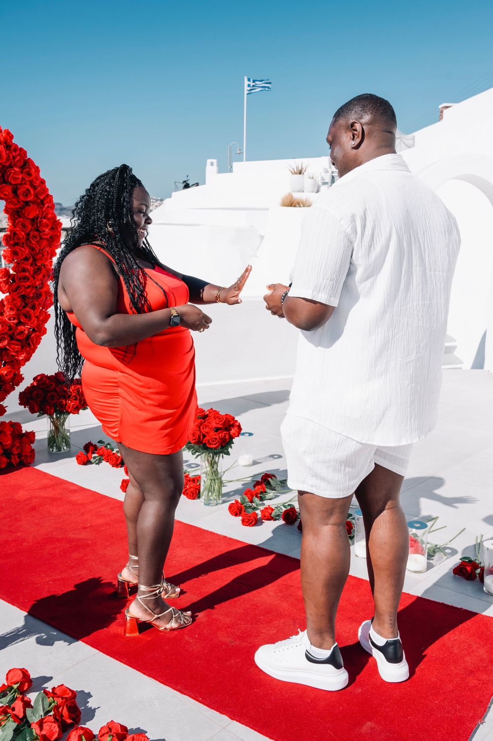 A couple celebrates their engagement moment on a red carpet under a floral arch.