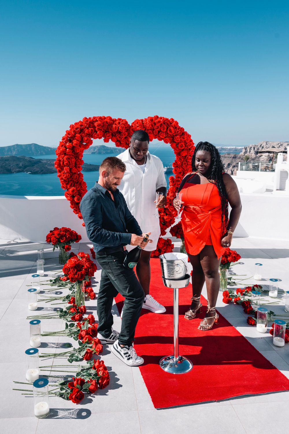 Romantic proposal setup with red heart arch and rose petals on white terrace overlooking Mediterranean Sea.