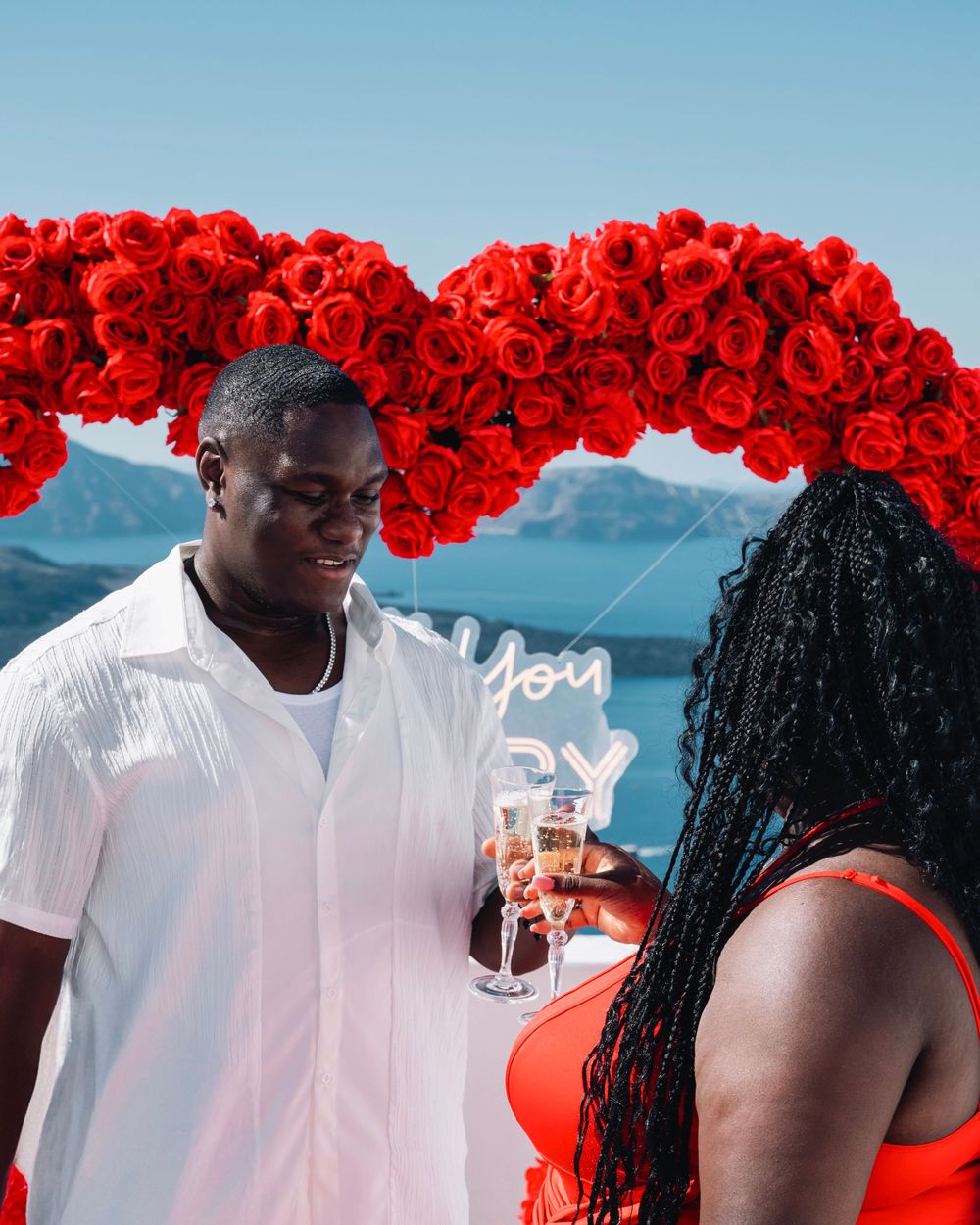 Couple celebrating with champagne under red rose heart arch with ocean view backdrop.