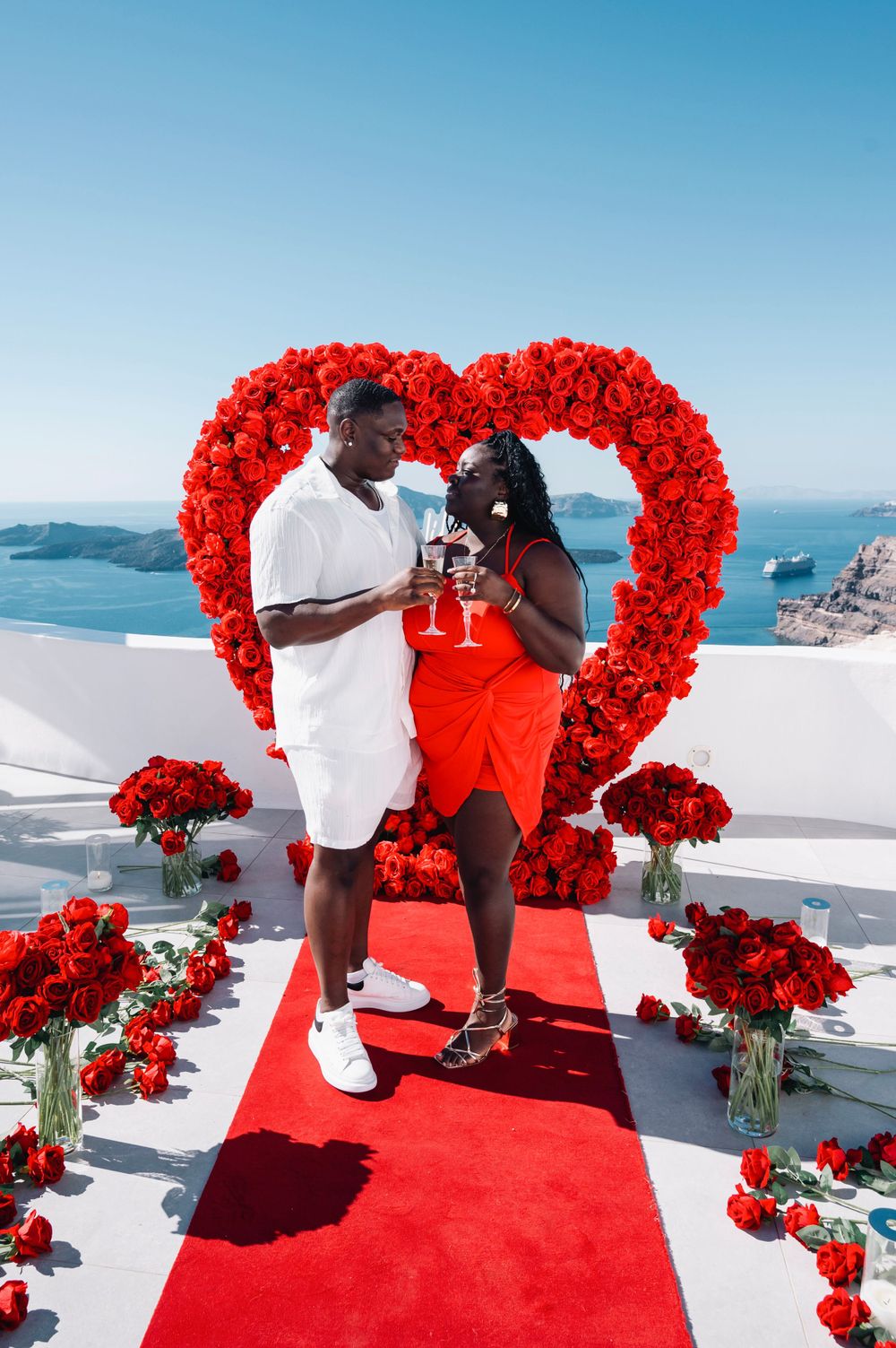Couple sharing romantic moment on red carpet under floral heart arch with ocean view.