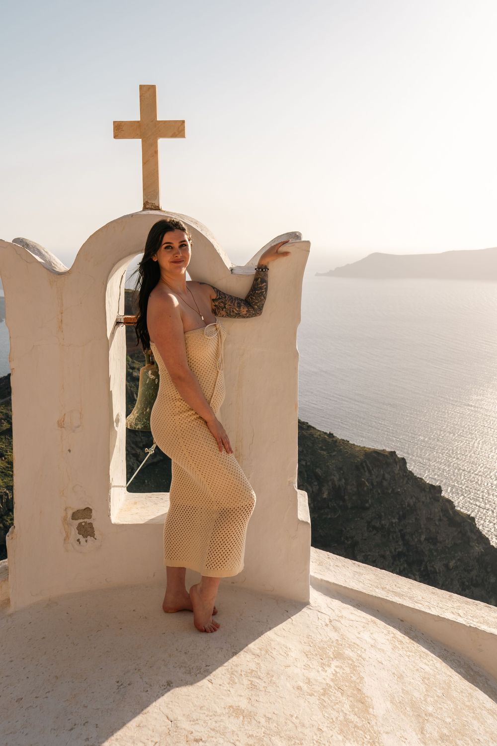 Woman in yellow dress posing by white arch with cross, overlooking Santorini caldera at sunset.