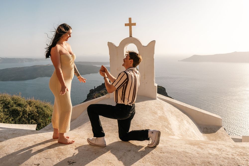 Man proposing to woman on one knee in Santorini, Greece, with scenic caldera views and white chapel cross backdrop.