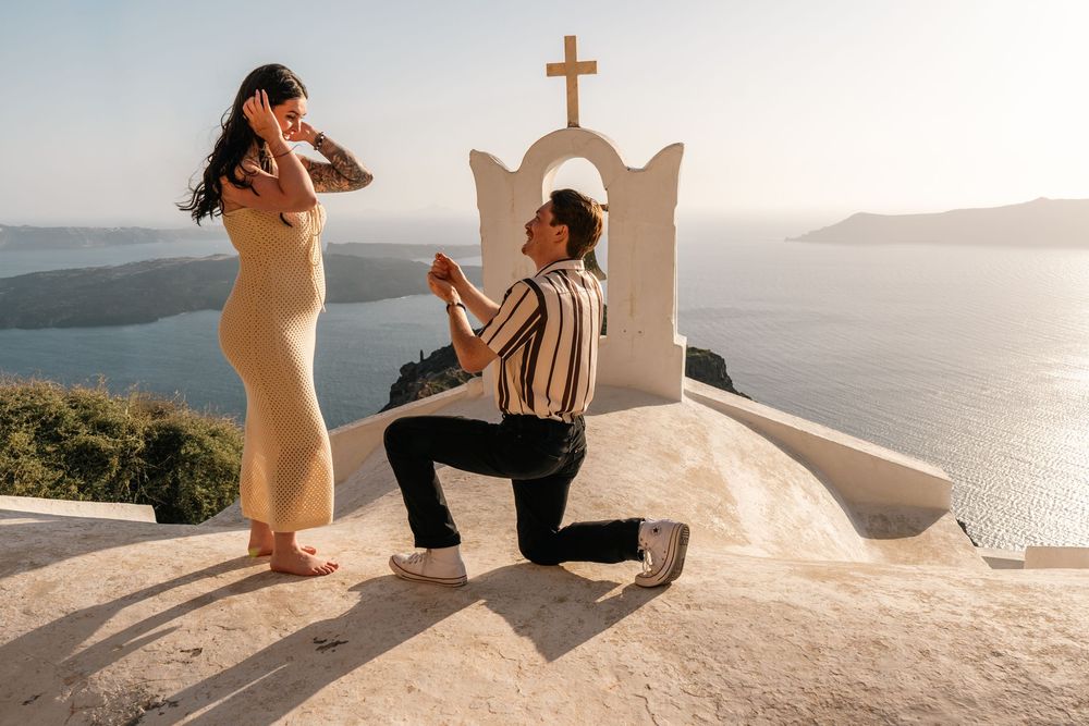 Man proposing to woman on one knee at Santorini chapel with stunning Aegean Sea views at sunset.
