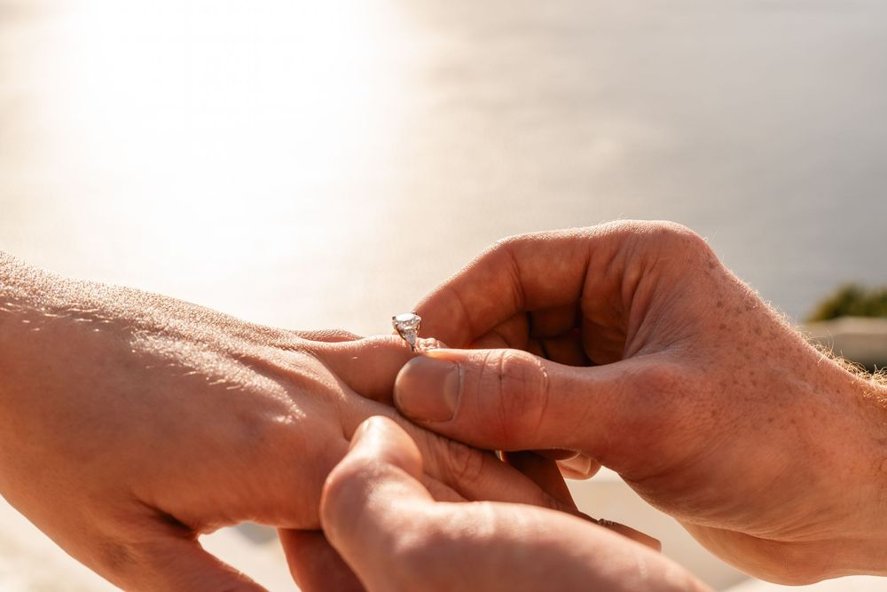 Close-up of a person placing a diamond engagement ring on their partner's finger outdoors in sunlight.