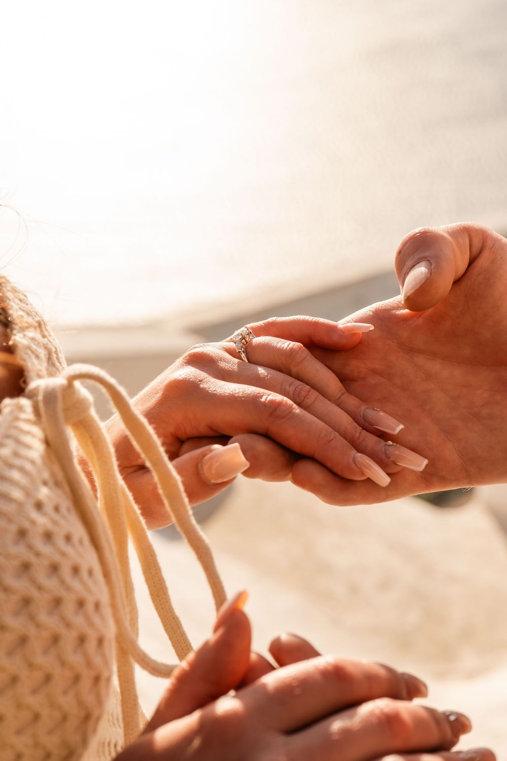Close-up of couple holding hands on beach, woman wearing engagement ring, sunny day.