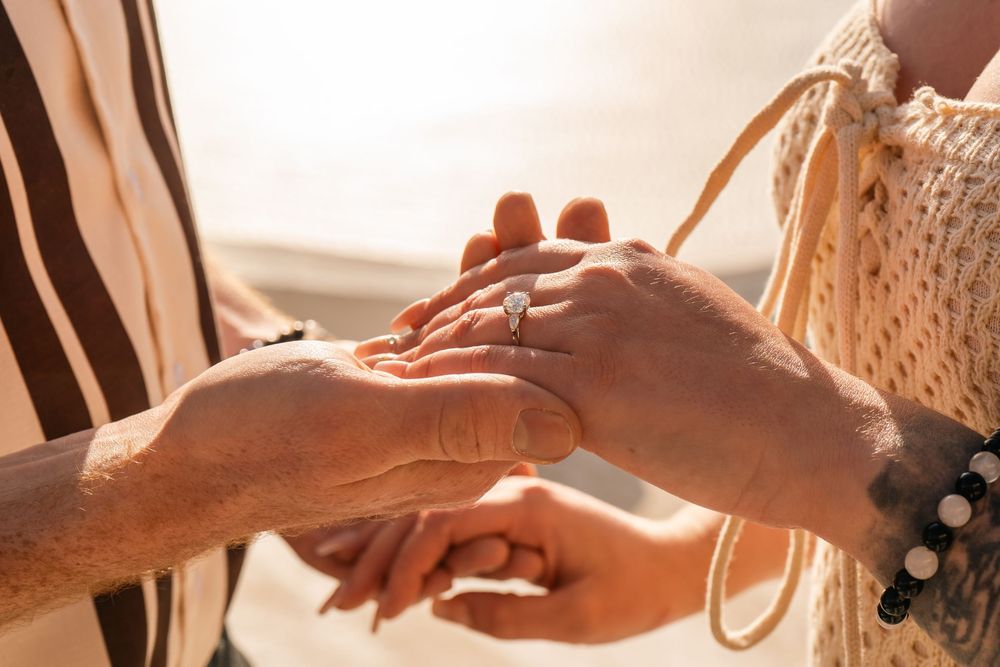 Close-up of elderly couple holding hands with diamond engagement ring on beach at sunset.