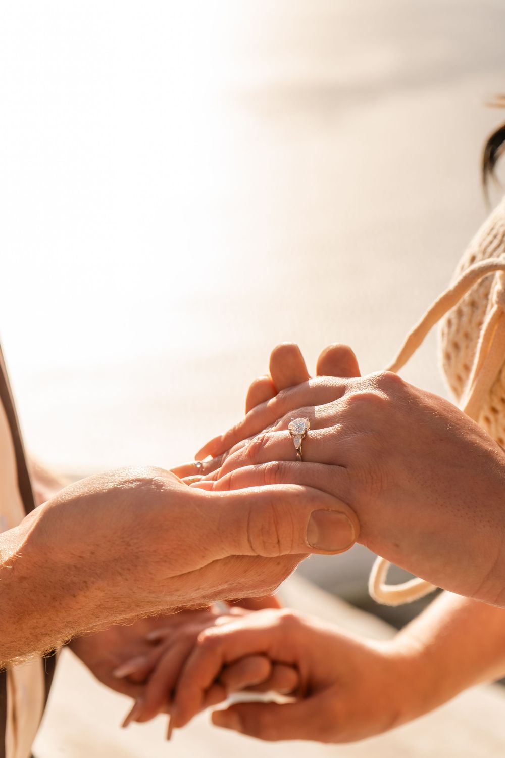 Close-up of a couple's hands as a man places a diamond engagement ring on a woman's finger at a sunny beach.